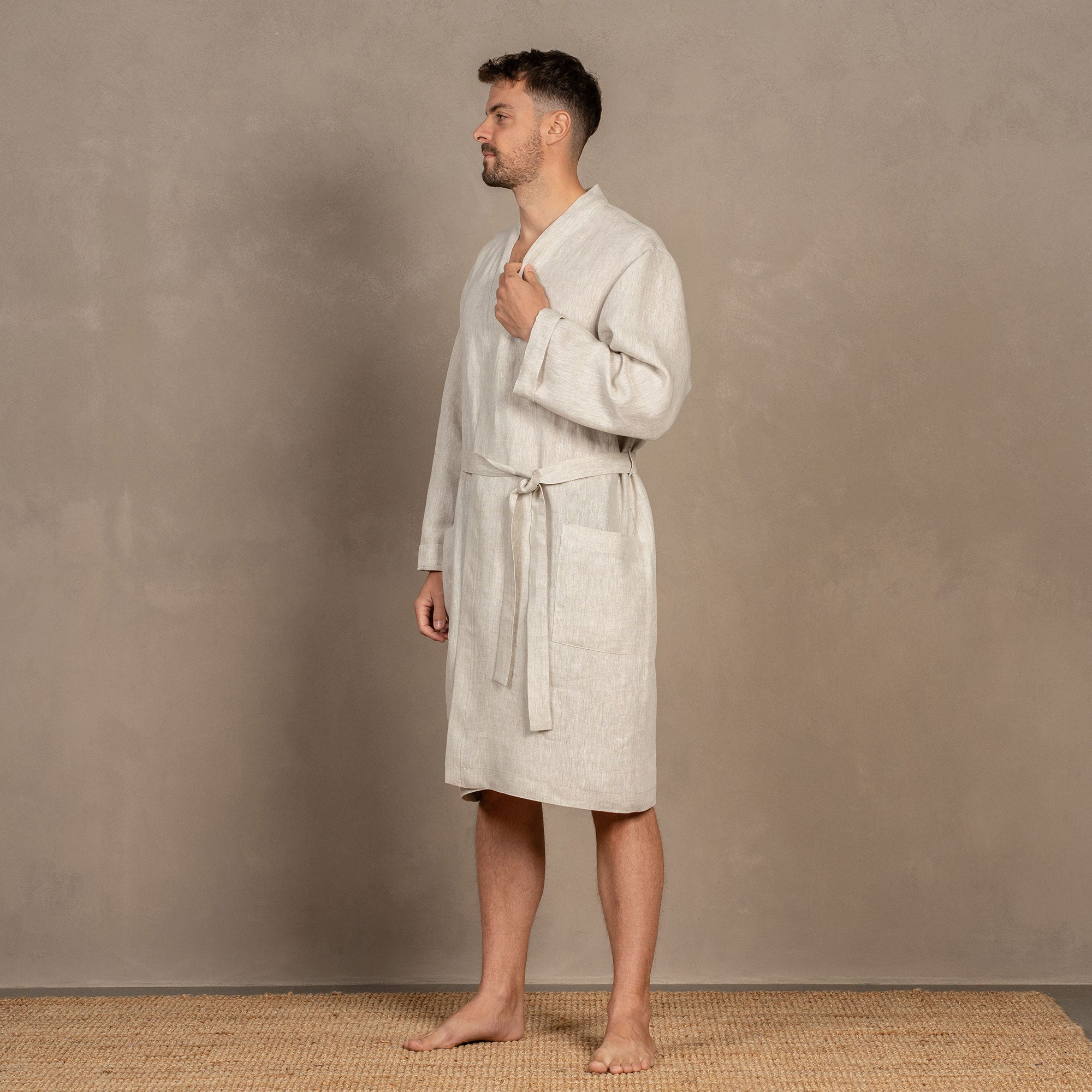 A man, turned slightly to the side, wearing the men's linen bathrobe in natural color. Both of his hands are in the side pockets.