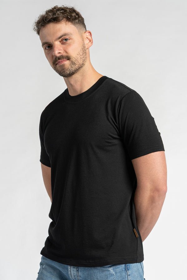 A man wearing a merino wool black t-shirt and blue jeans is shown from the chest up against a light background.  He has short brown hair and a light beard, and is looking slightly to the side. The menique black t-shirt is the primary focus, showcasing its fit and appearance.