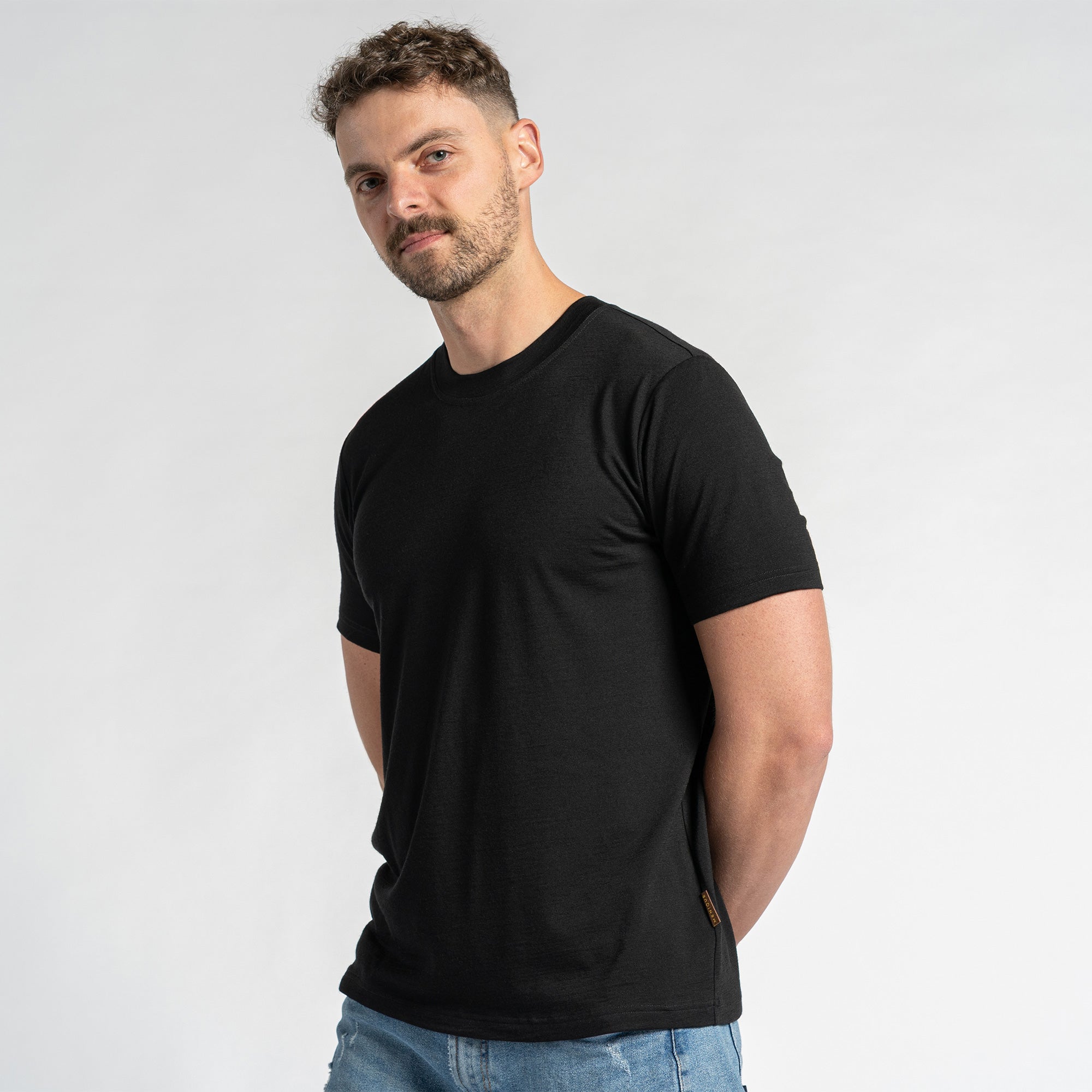 A man wearing a merino wool black t-shirt and blue jeans is shown from the chest up against a light background.  He has short brown hair and a light beard, and is looking slightly to the side. The menique black t-shirt is the primary focus, showcasing its fit and appearance.