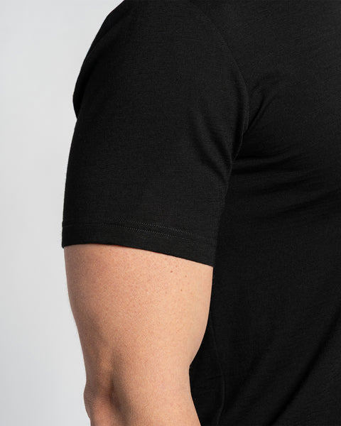 Close-up of a merino black t-shirt sleeve, showing the fabric texture and seam details. The t-shirt is displayed on a person&#39;s arm, with a light background.