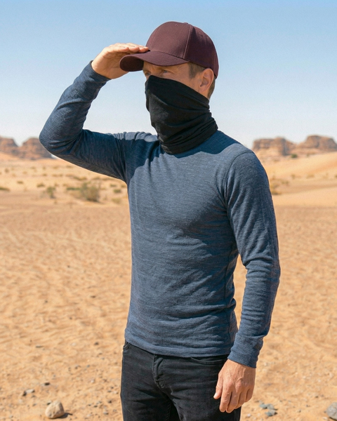 A man standing in a desert landscape, turned slightly to the side in a three-quarter profile view. He is wearing the same outfit: a dark maroon baseball cap, a black neck gaiter covering his lower face, a blue long-sleeved shirt, and black pants. He has his right hand raised to the brim of his cap, shielding his eyes from the sun as he looks out into the distance.