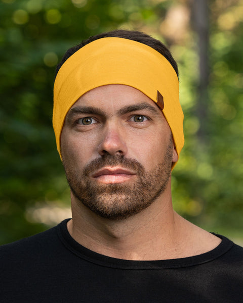 A close-up of a man&#39;s face shows him wearing a spicy yellow menique headband with a small brown tag. He has short, dark hair, light eyes, and a short beard. He is wearing a black shirt and looking directly at the camera with a serious expression. The background is softly blurred green and brown, suggesting an outdoor setting.