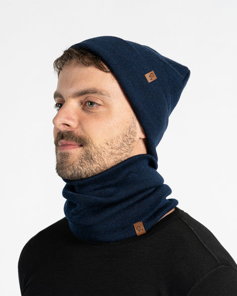 A man with a light brown beard is shown from the chest up, looking to the left of the frame. He is wearing a blue knitted beanie and a matching neck gaiter. Both items of clothing have a small brown leather patch with a menique logo on them. The background is a plain white.