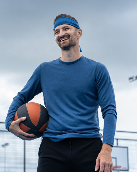 A smiling man stands on an outdoor basketball court holding a basketball under his right arm. He is wearing a blue long-sleeve athletic shirt, a matching blue tie headband, and black athletic pants. A chain-link fence and a basketball hoop are visible in the background beneath a cloudy sky.