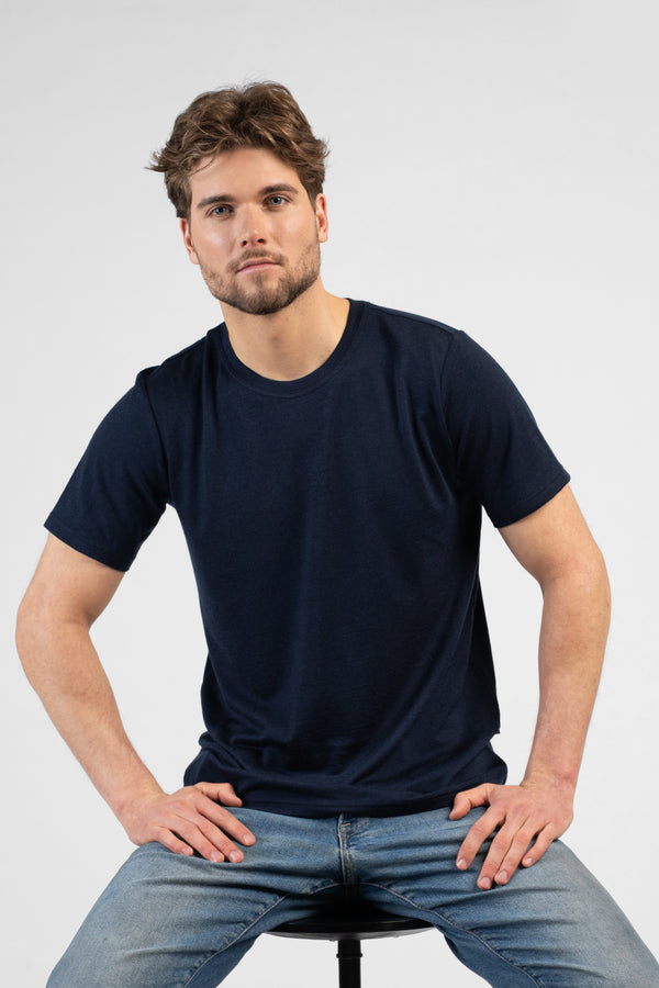 A studio portrait of a man sitting on a black stool against a plain white background. He is leaning slightly forward with his hands resting on his upper thighs, looking directly at the camera. He is wearing a dark navy blue short-sleeved crew-neck t-shirt and blue jeans.