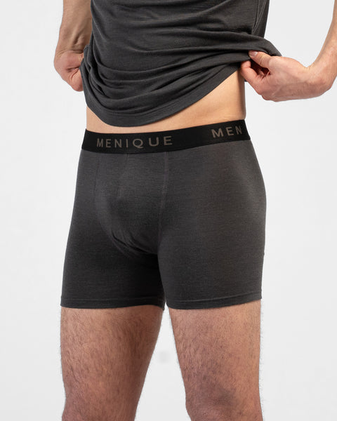 A close-up lifestyle shot of a man's torso and thighs against a plain white background. He is lifting the hem of a charcoal grey t-shirt with both hands to reveal matching charcoal grey short boxer briefs. The underwear features a contrasting black elastic waistband with "MENIQUE" woven in light brown text.