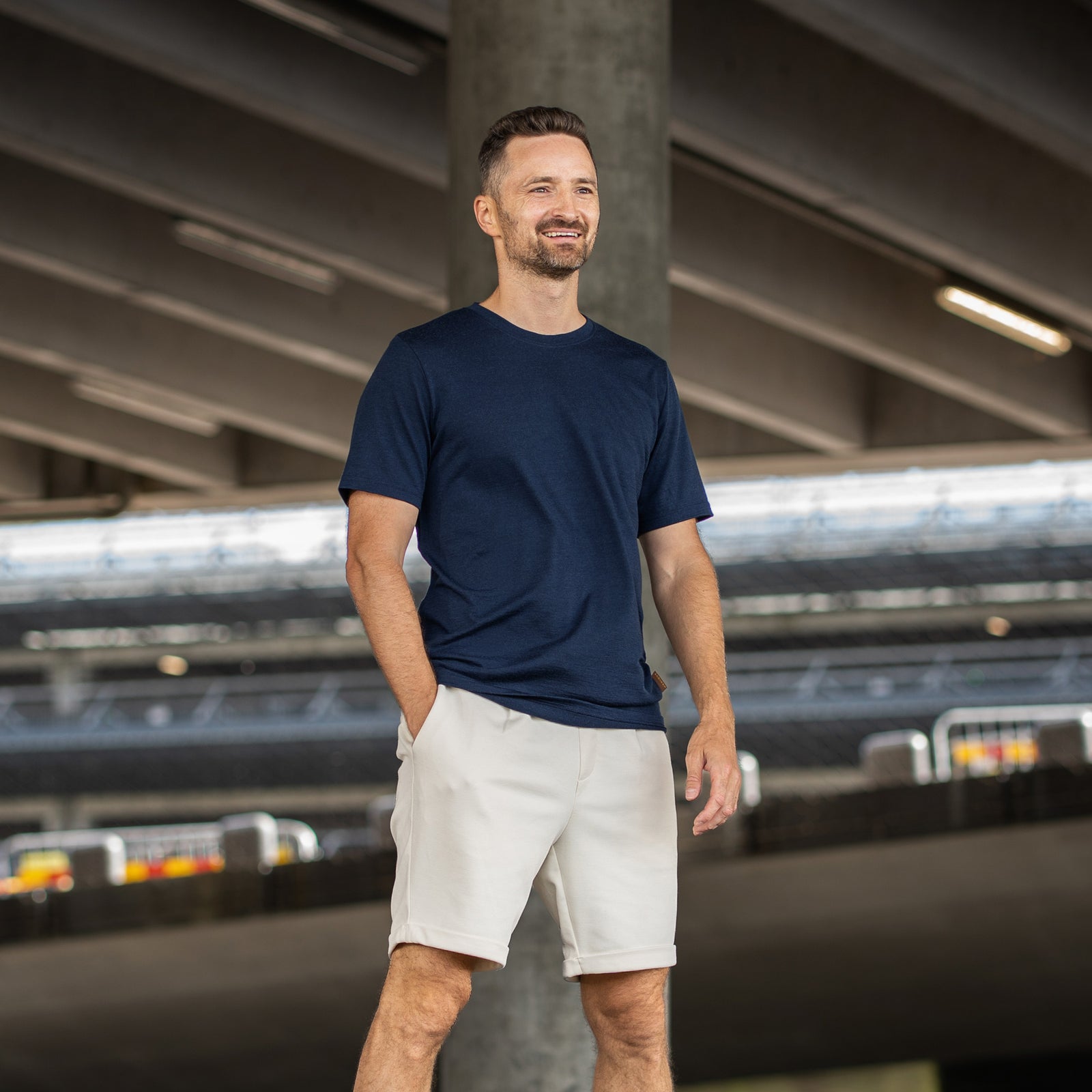 A smiling man standing outdoors in front of rusty metal pillars. He is wearing a navy blue short-sleeve crew neck t-shirt and beige shorts, with his right hand casually resting in his pocket as he looks off into the distance.