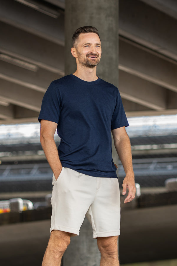 A smiling man with a short beard stands looking off to the right, with one hand resting in his pocket. He is wearing a navy blue short-sleeve crew neck t-shirt and light beige shorts. The background features the shaded underside of a large concrete structure or overpass.