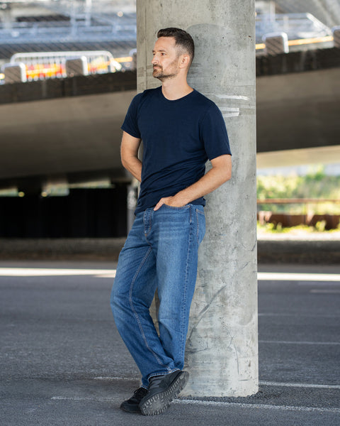 A smiling man stands outdoors with his right hand in his pocket, looking toward the right. He is wearing a navy blue short-sleeve crew neck t-shirt and light beige shorts. He is positioned in front of a textured, rusted metal barrier.
