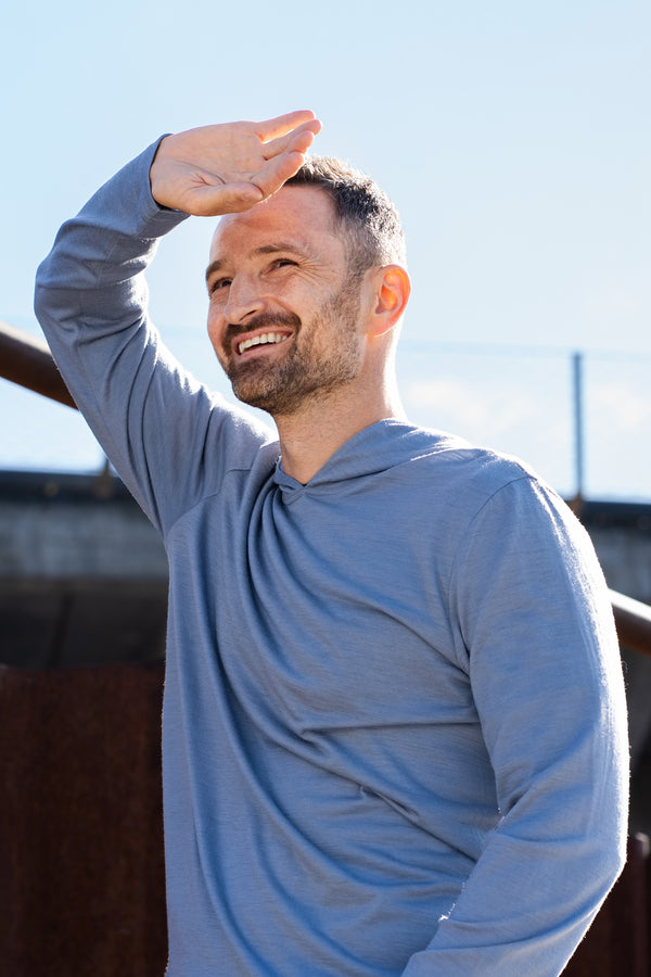 A smiling man stands outdoors on a sunny day, shielding his eyes from the sun with one hand. He is wearing a long-sleeve, dusty blue hoodie. In the background, there is a rusty metal structure and part of an elevated concrete overpass.