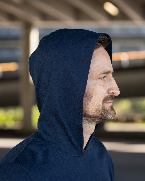 A close-up profile shot of a man's face and upper body outdoors. He is looking off to the right, wearing a navy blue hoodie with the hood pulled up over his head. The background is a blurred concrete structure, likely under a bridge.