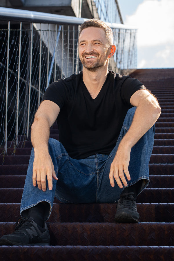 A smiling man sitting on outdoor metal stairs. He is wearing a short-sleeve black V-neck t-shirt, loose blue jeans, and black sneakers, resting his hands casually on his knees.