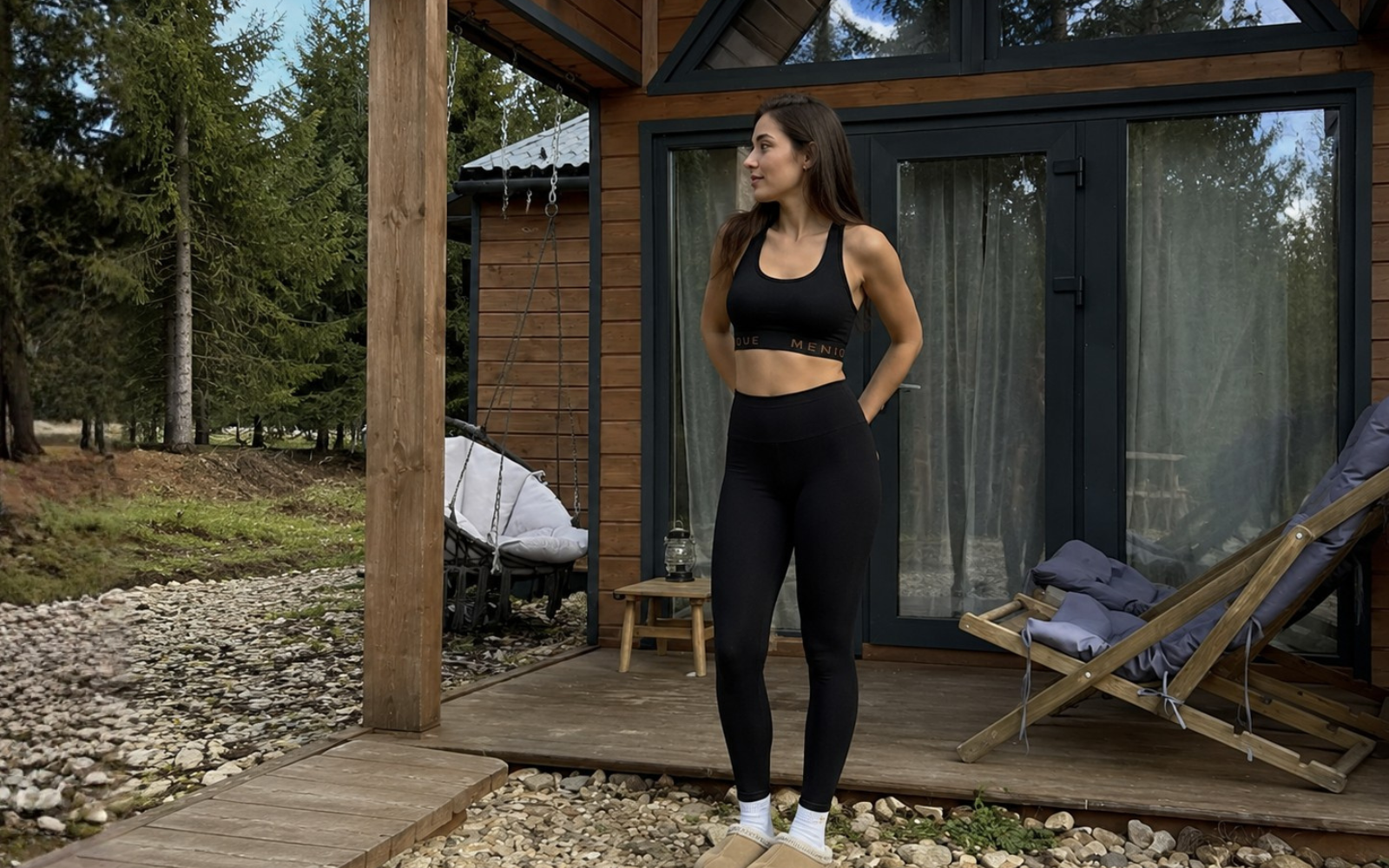 A woman stands on a wooden deck outside a cabin wearing a black sports bra and matching black leggings.