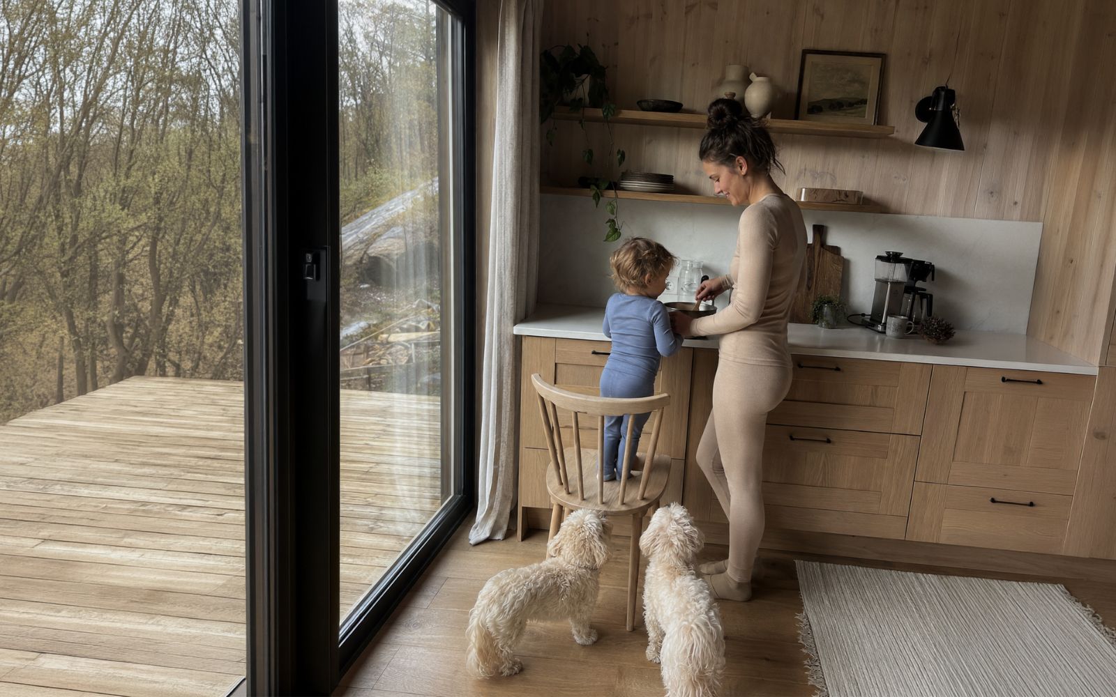A woman in beige activewear stands in a modern wood kitchen while a child in blue stands on a wooden chair at the counter. Two small white dogs are on the floor in the foreground, and a large window looks out onto a wooded outdoor deck.