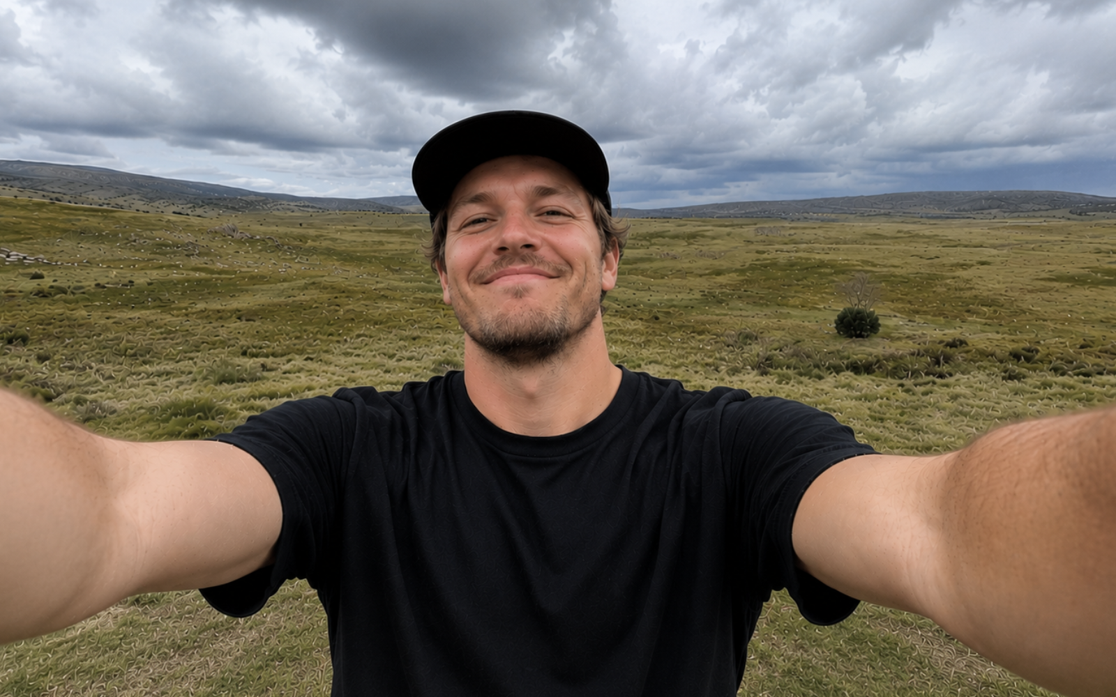 A man taking a selfie outdoors in a grassy landscape under a cloudy sky, wearing a black t-shirt and black cap.