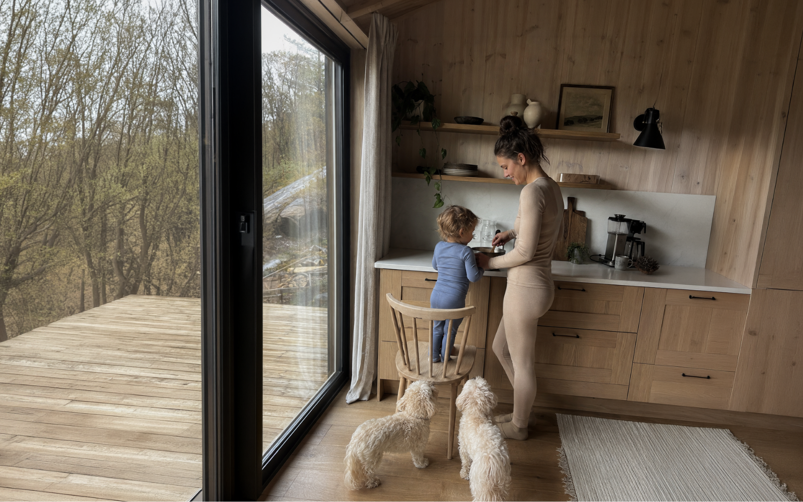 A woman and a young child, both wearing light-colored matching sets, standing by a kitchen counter next to large windows looking out onto a wooded area.