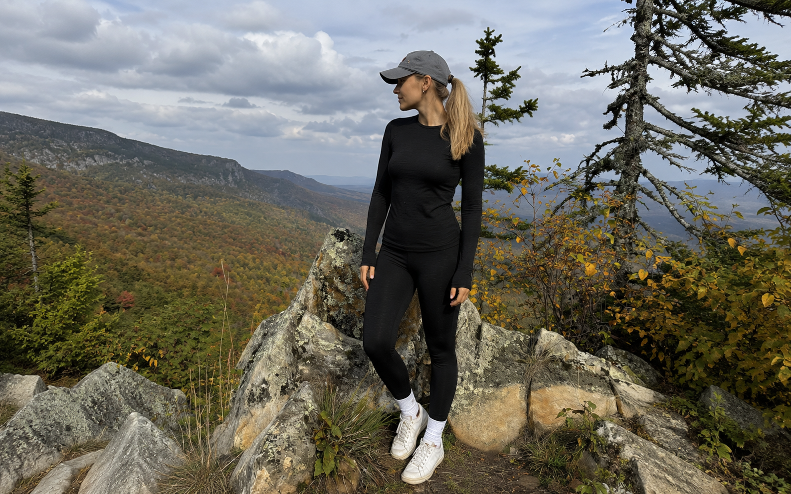 A woman stands on a rocky mountain overlook wearing a black long-sleeve athletic top and black leggings with white sneakers and a gray baseball cap, with a mountain valley and trees in the background.