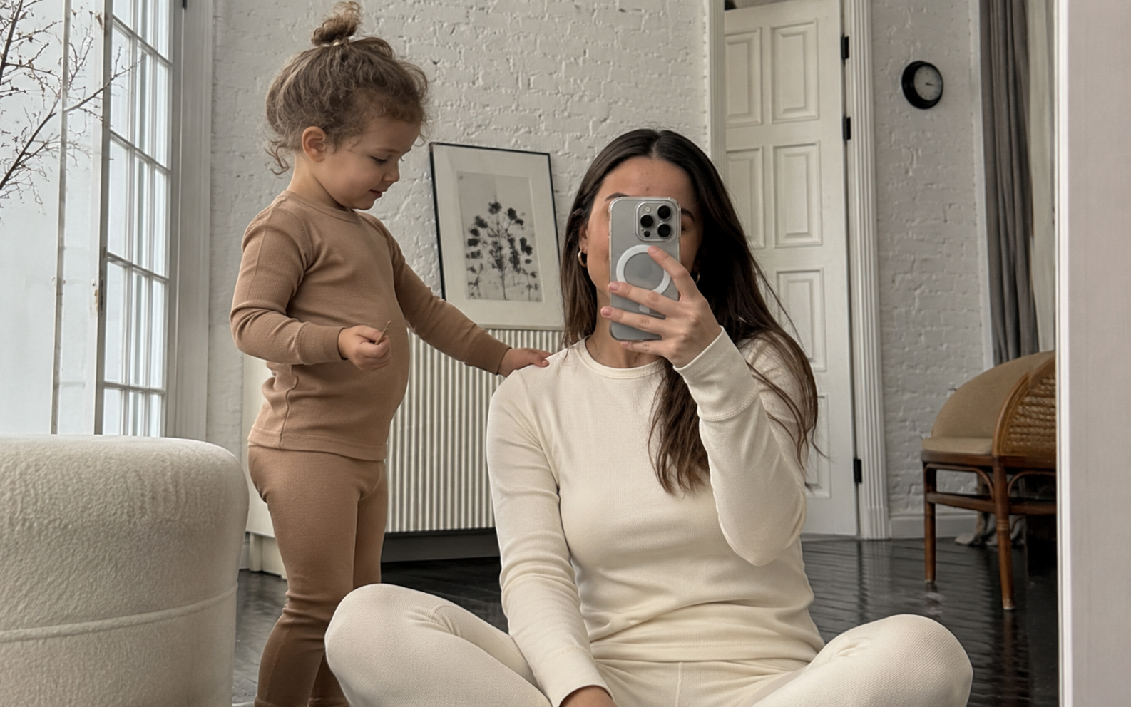 A woman and a young child wearing matching beige and cream loungewear sets in a bright, modern living room. The woman is sitting on the floor taking a mirror selfie, while the child stands beside her.