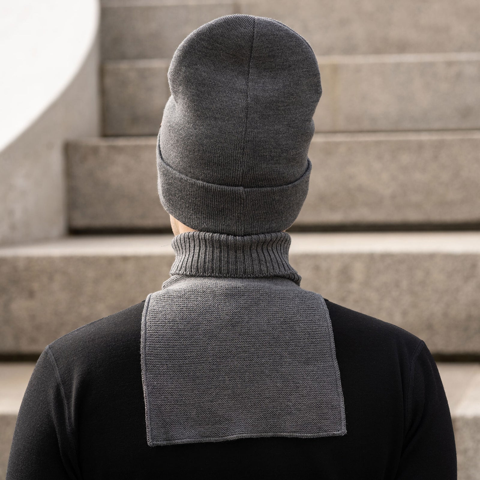A man shown from the chest up, turned slightly to his right and smiling. He is wearing a dark grey knit beanie with a small brown logo patch and a matching dark grey knit neck warmer over a black long-sleeve shirt. The background consists of blurred concrete stairs.