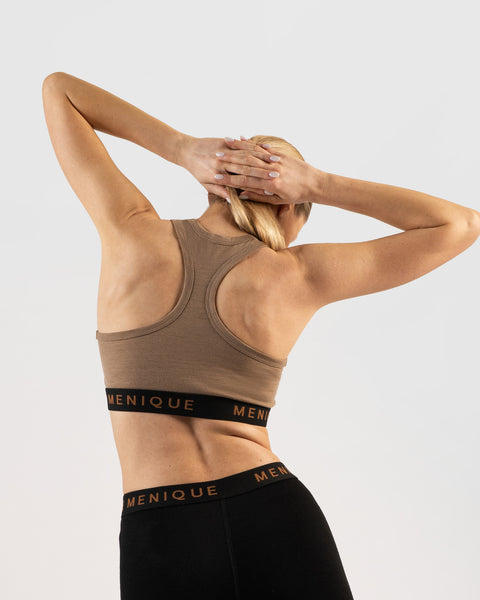 A rear-view studio portrait of a woman's back and shoulders. She has her hands clasped together behind her head, elbows pointing outward. She is wearing a beige racerback sports bralette with a black elastic band featuring "MENIQUE" in light brown text, and matching black athletic leggings. The background is a solid light grey.