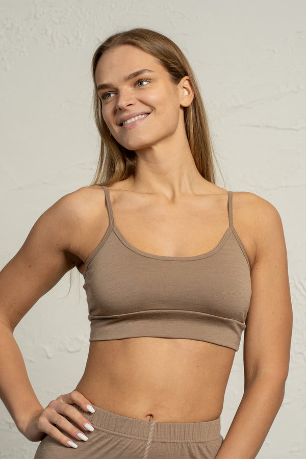 A woman wearing a beige merino bralette smiles softly while posing with one hand on her hip. The bralette has thin adjustable straps and a smooth, fitted design. She stands in front of a light textured background.