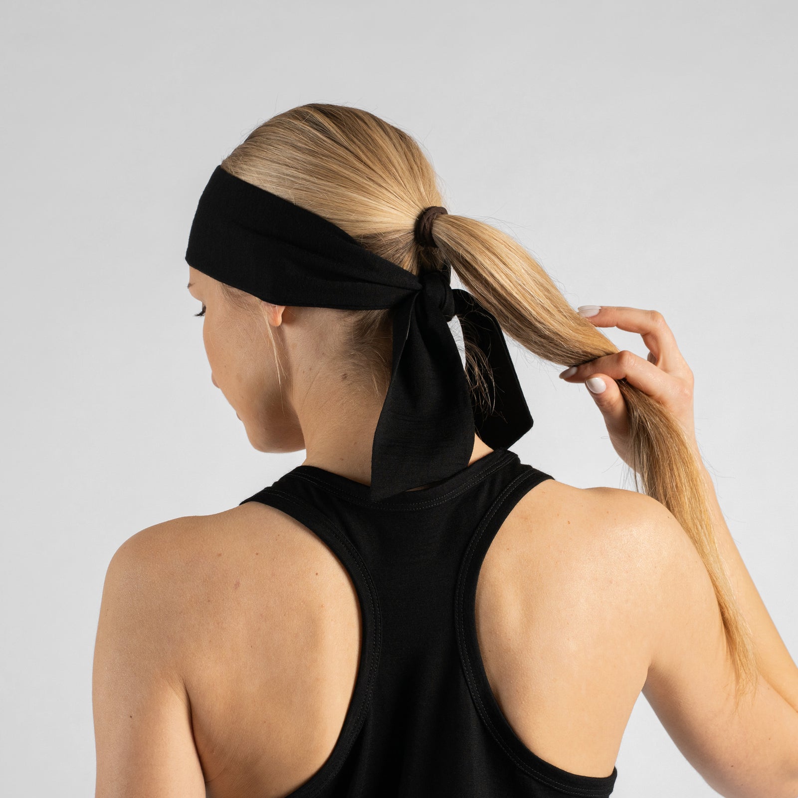 A studio portrait of a woman looking downwards and to the right as she ties a solid black headband behind her head. Her blonde hair is pulled back into a ponytail, and she is wearing a black tank top. The background is a solid light grey.