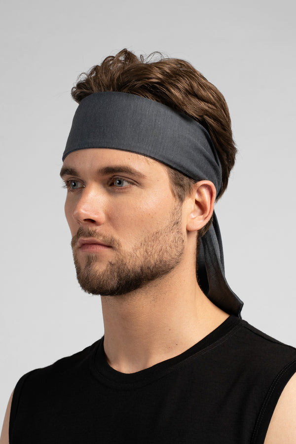 A studio portrait of a man looking thoughtfully off to the left side of the frame. He is wearing a medium grey tie-back headband, with the tied ends hanging down behind his neck. He is wearing a black sleeveless top against a solid light grey background.