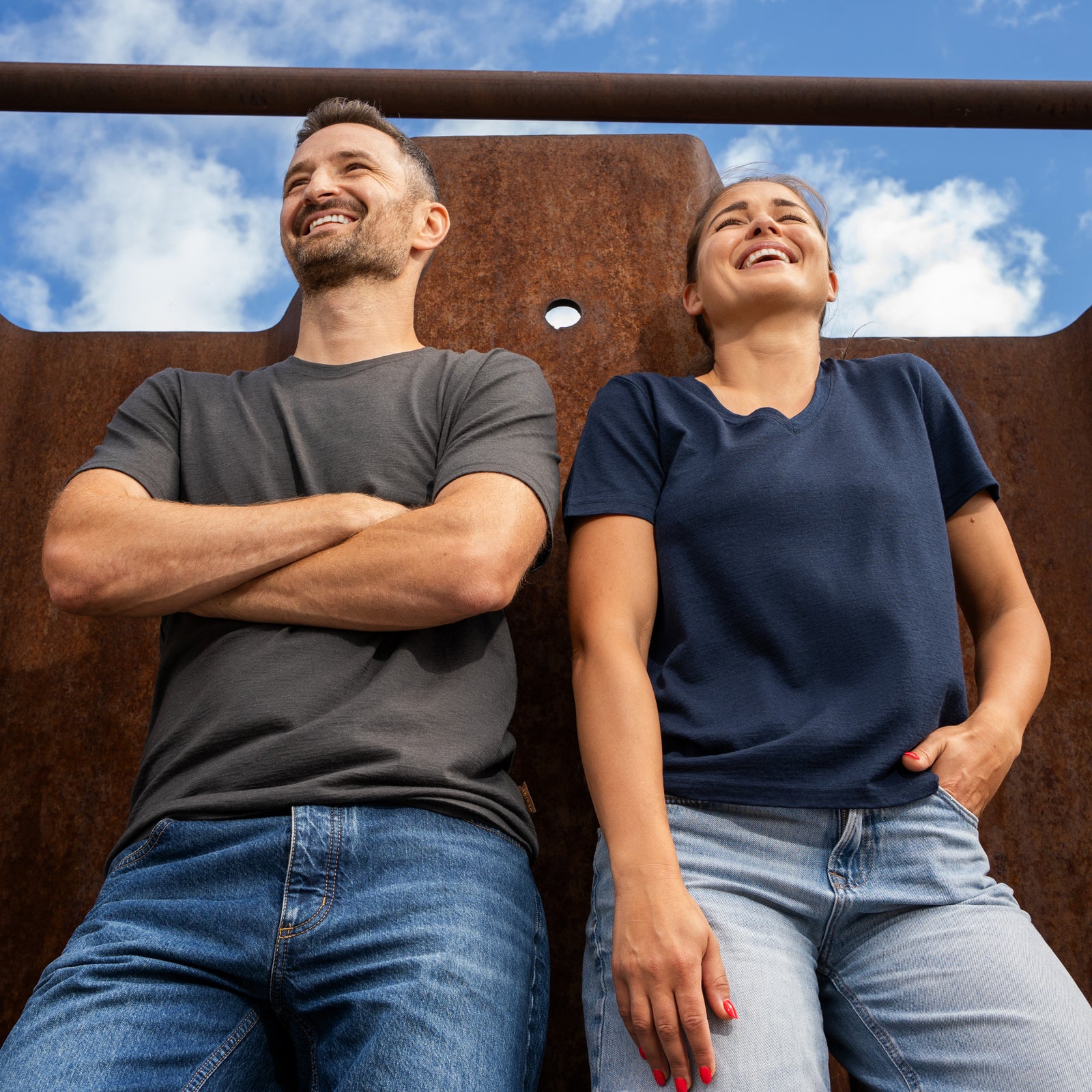 A woman standing next to a textured, rusty metal wall, smiling and looking away from the camera. She is wearing a navy blue v-neck t-shirt and blue jeans, with her right hand resting gently on the metal structure and her left hand in her pocket.