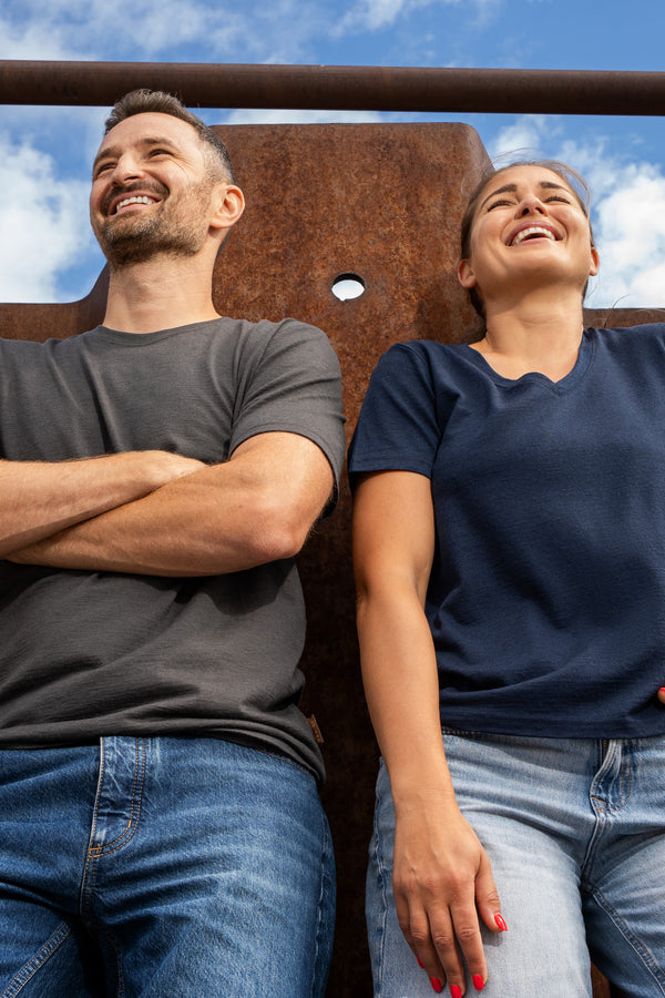 A low-angle shot of a smiling man and a laughing woman leaning back against a large, rusted metal wall under a blue sky with clouds. The man is on the left with his arms crossed, wearing a dark grey short-sleeve t-shirt and blue jeans. The woman is on the right with one hand resting in her pocket, wearing a navy blue short-sleeve v-neck t-shirt and light blue jeans.