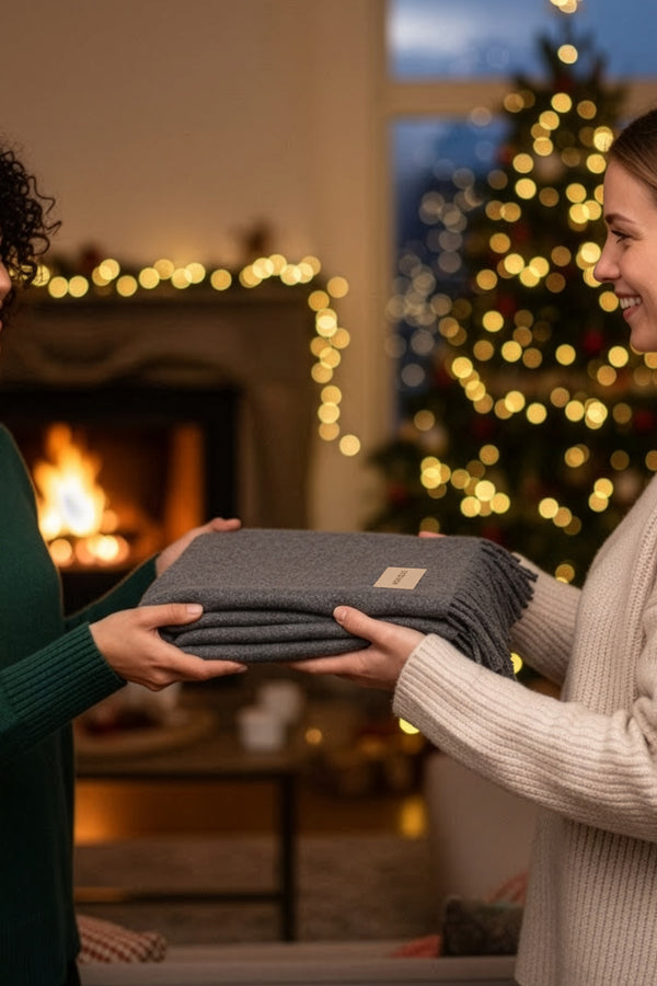 Two women exchanging a folded blanket in a cozy living room with a Christmas tree and fireplace.