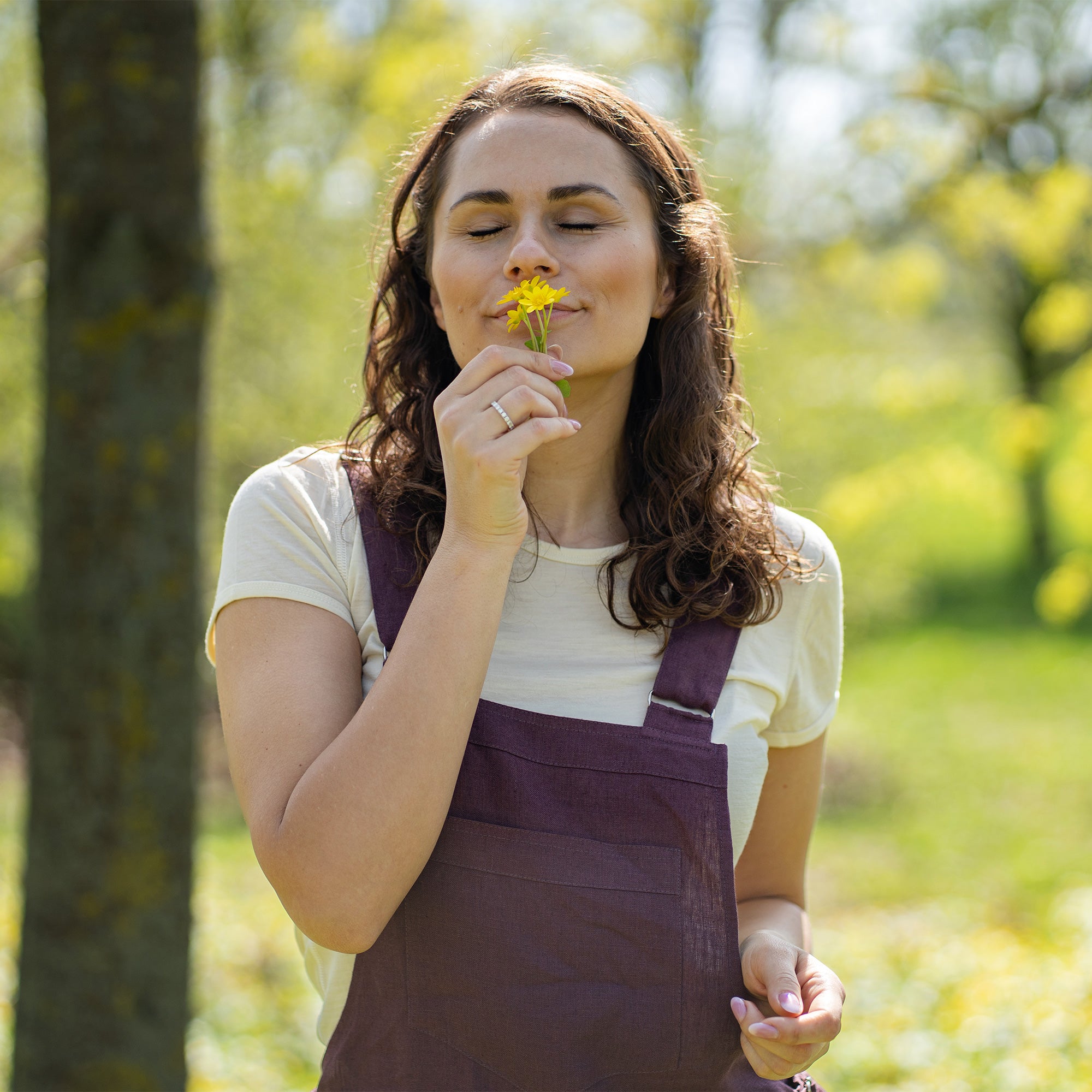 A woman with brown hair, wearing dark purple overalls over a light-colored t-shirt, stands outdoors between two tree trunks on grassy ground.
