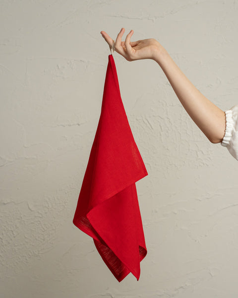 A person’s hand holding up a red linen kitchen towel against a textured light wall.