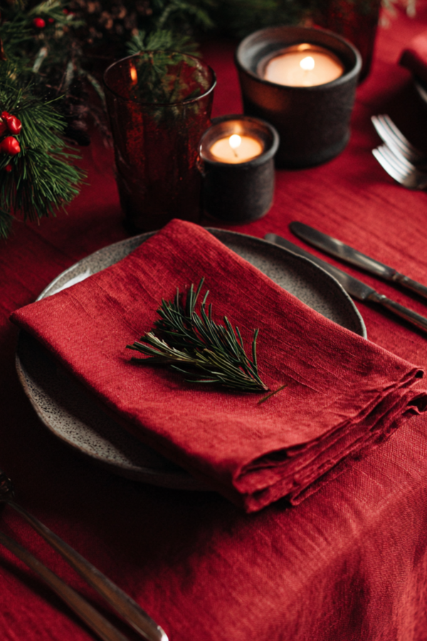 Festive Christmas table setting with a red linen napkin folded on a stone plate, decorated with a pine sprig and surrounded by candles and greenery.