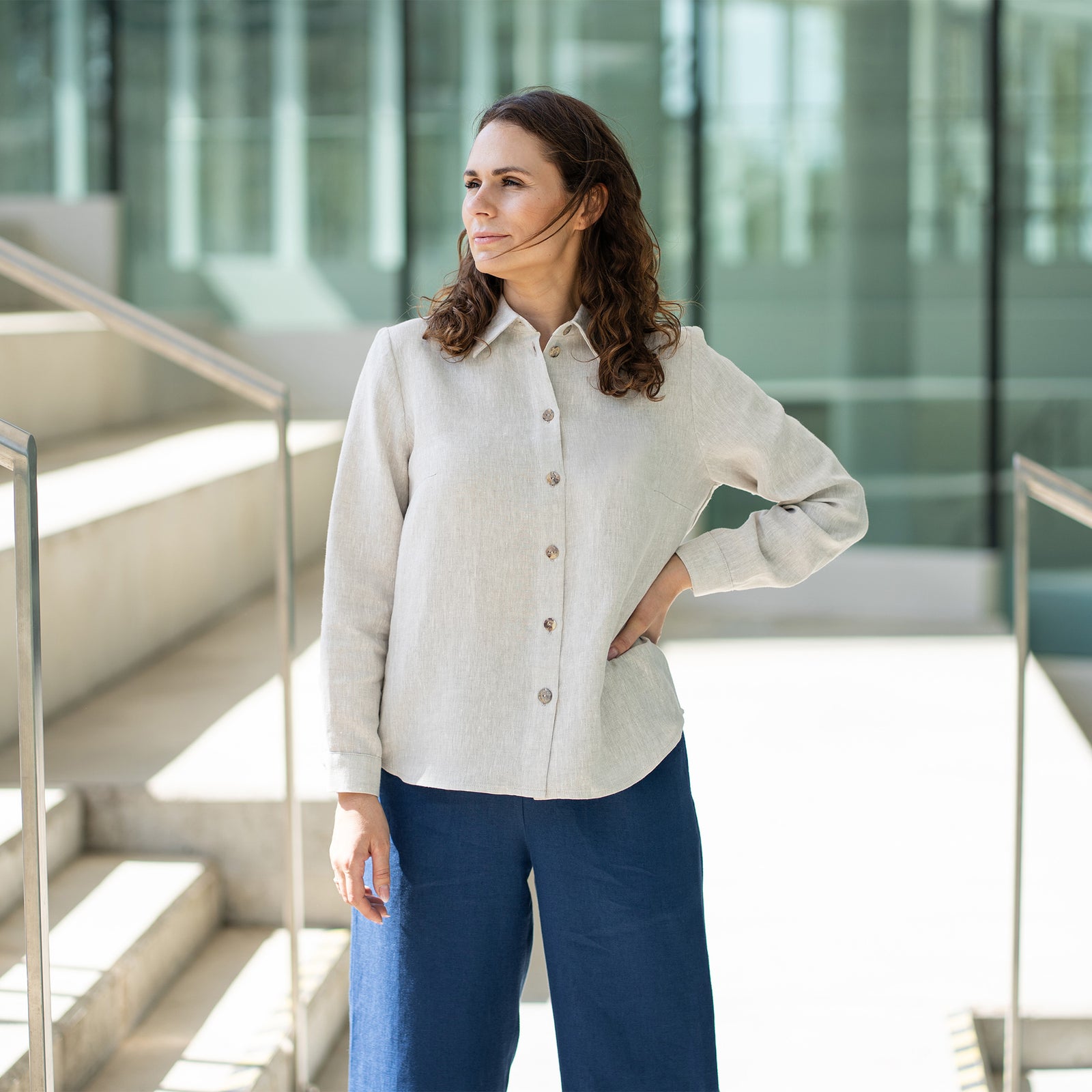 A woman with brown hair, wearing a light blue button-front shirt and wide-leg white trousers, stands outdoors against a modern concrete structure.

