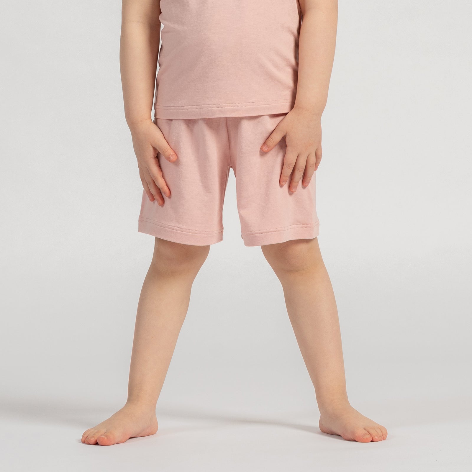 A studio shot, cropped from the chest down, of a young child standing barefoot. The child has their hands resting on their hips. They are wearing a matching set of a light sage green short-sleeved t-shirt and shorts. The background is a plain light grey.