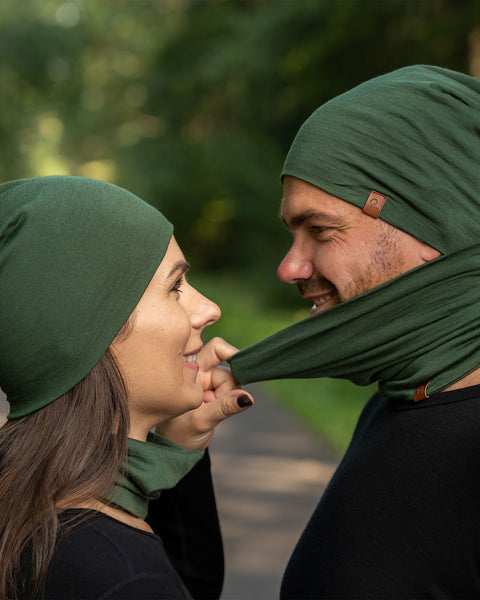 The picture features a man and a woman wearing the unisex dark green merino gaiter and beanie set. They are in a forest. The woman is pulling the man&#39;s gaiter to remove it. Both of them are smiling. 