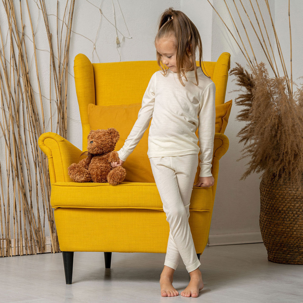 a boy sitting on a bed, wearing the Kids' Merino 160 Long Sleeve Set in Natural color.