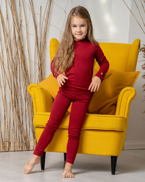 A girl, standing by a yellow armchair, wearing the menique Kids&#39; 160 Merino Pants in Royal Cherry color, with one leg slightly to the side and both hands on hips. 