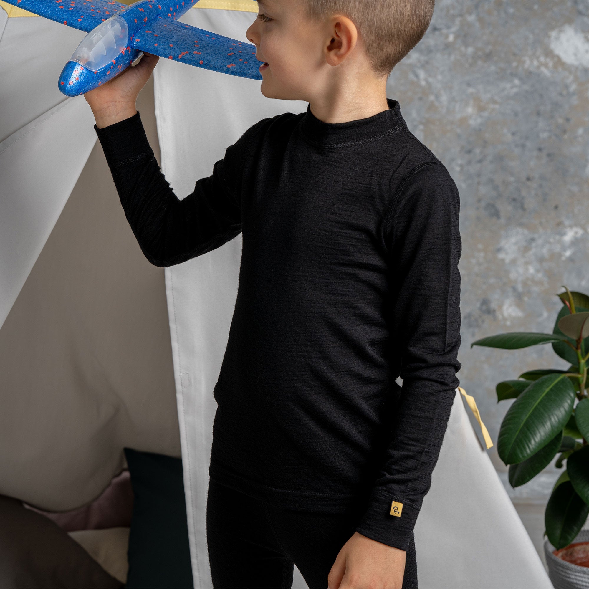 A young child with blonde hair and blue eyes, wearing a black long-sleeved shirt and blue jeans, is hanging onto a metal bar on a playground structure. They are looking directly at the camera with a happy, open-mouthed expression and their tongue slightly sticking out.