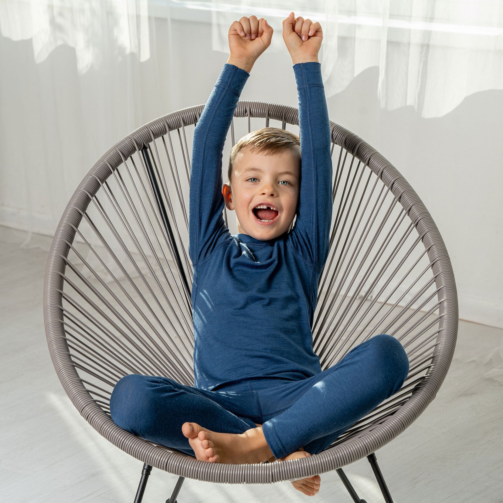 A boy sitting in a lounge chair, wearing the menique Kids' Merino 160 Long Sleeve Set in Denim color. 