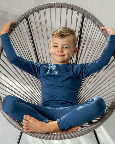 A boy wearing the menique kids&#39; 160gsm merino long sleeve set in denim color. he is sitting in a lounge chair, with his legs crossed and hands raised.