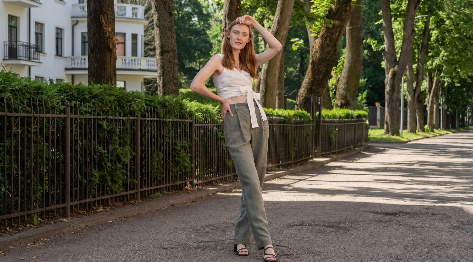 A woman with long red hair stands on a quiet street lined with trees, wearing a white wrap-style linen top tied at the waist and high-waisted olive green linen trousers. She poses confidently with one hand resting on her head, dressed in minimalist sandals, with soft sunlight highlighting the natural textures of the outfit.