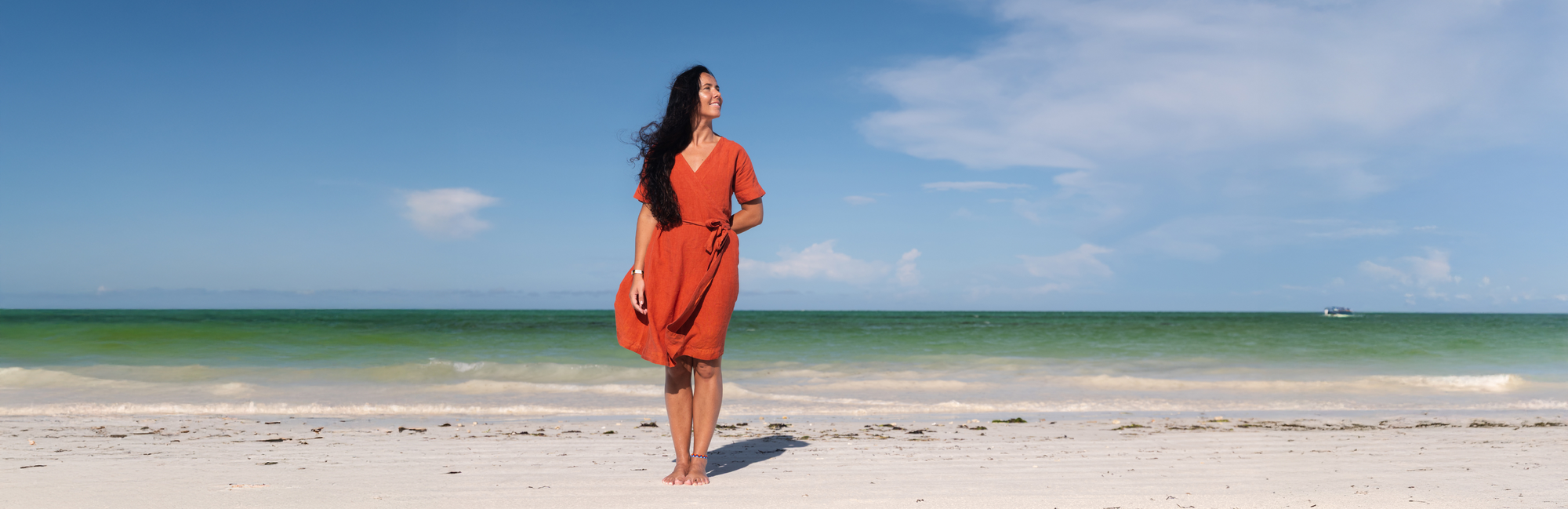 A woman with long dark hair stands on a sandy beach, facing away from the camera. She wears a long-sleeved orange dress and is barefoot. The ocean is in the background with light waves, and the sky is blue with a few clouds.