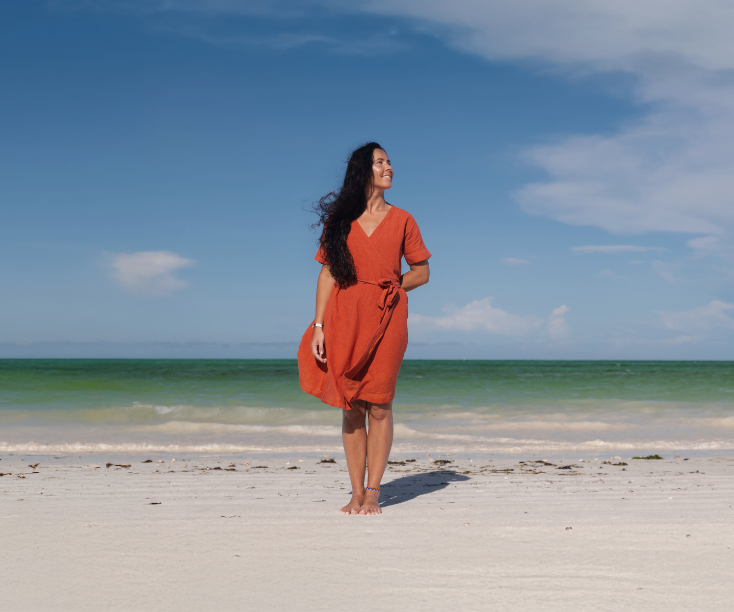 A woman with long dark hair stands on a sandy beach, facing away from the camera. She wears a long-sleeved orange dress and is barefoot. The ocean is in the background with light waves, and the sky is blue with a few clouds.