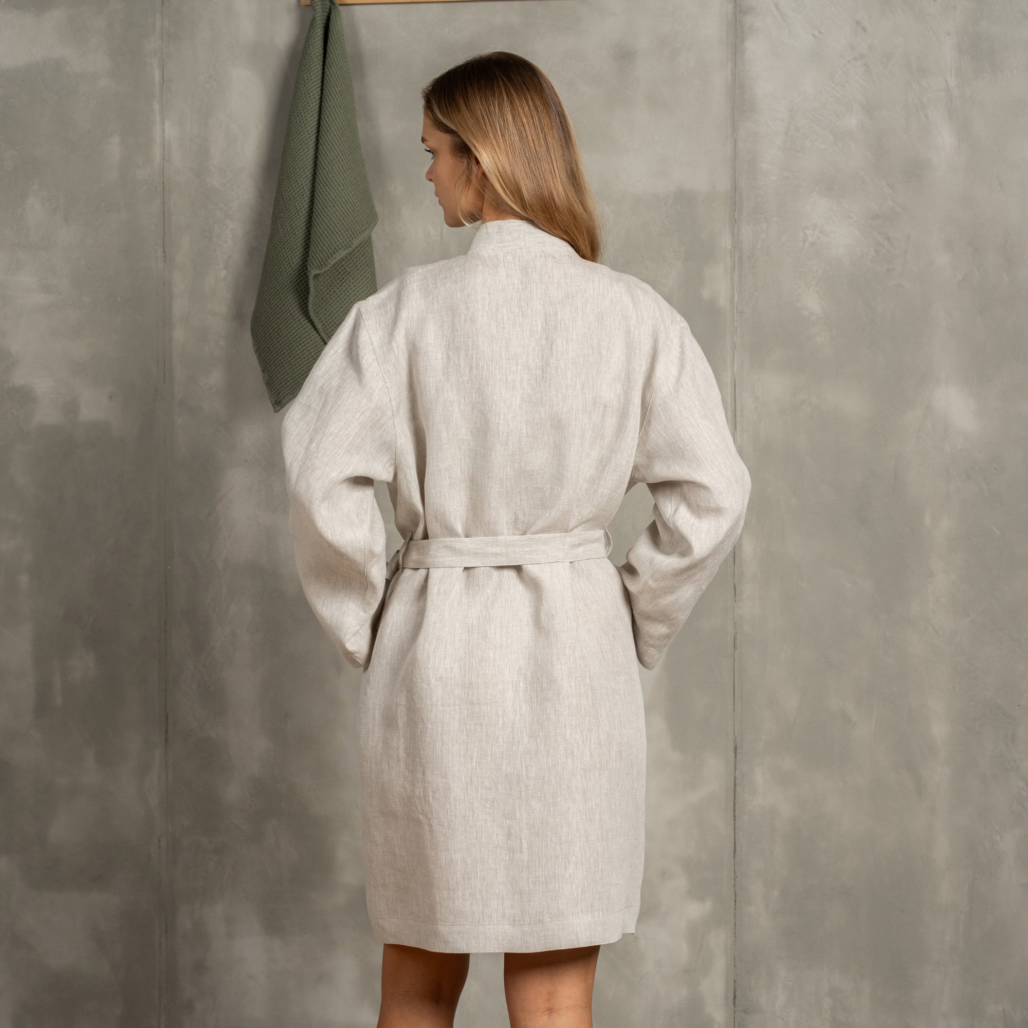 The menique women's linen bathrobe in natural color. The woman is standing in front of a gray wall with a wooden hanger on it. There is a linen waffle towel on the hanger. She has in the pocket, the other put lightly on her stomach.