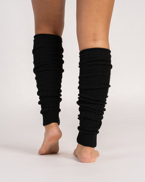 A view from behind of a person standing barefoot, wearing long, black, ribbed-knit leg warmers scrunched down their calves against a solid light gray background.