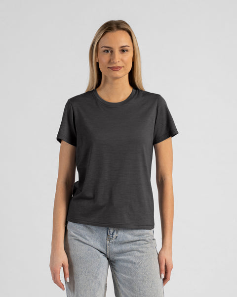 A studio portrait of a blonde woman standing and looking directly at the camera with her arms relaxed by her sides. She is wearing a regular fit, charcoal grey short-sleeved crew-neck t-shirt and light-wash blue jeans. The background is a solid light grey.