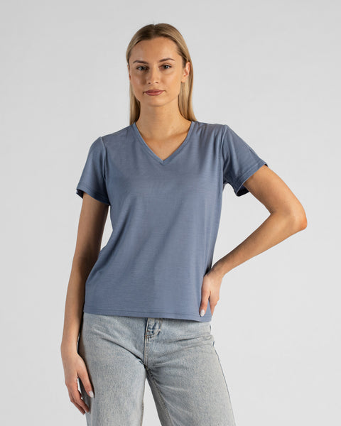 A studio portrait of a blonde woman standing and looking directly at the camera with a subtle smile. She is wearing a regular fit, dusty blue short-sleeved v-neck t-shirt and light-wash blue jeans. Her left hand is resting on her hip, while her right arm hangs relaxed by her side. The background is a solid light grey.