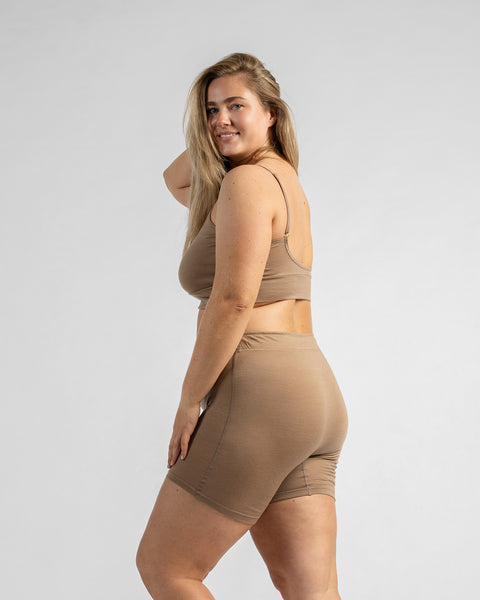 Side-back view of a model looking over her shoulder, wearing a beige sports bra and matching beige biker shorts.