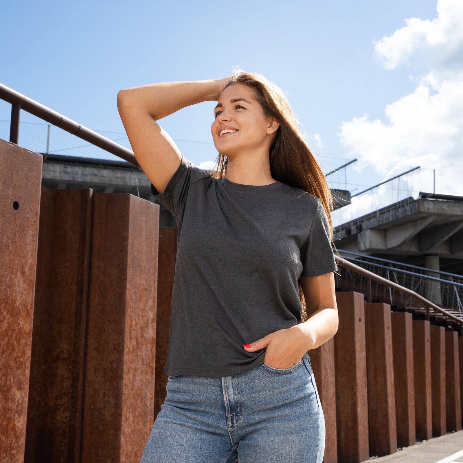 A studio portrait of a blonde woman standing and looking directly at the camera, with her left hand casually tucked into the front pocket of her light-wash blue jeans. She is wearing a regular fit, charcoal grey short-sleeved crew-neck t-shirt against a solid light grey background.